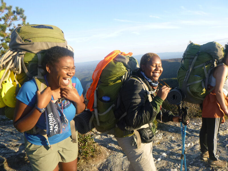 The image shows three women hiking with large backpacks. They are on a rocky terrain with a scenic view in the background. The woman in the front is laughing and wearing a blue shirt and khaki shorts. The other two women are also smiling, and they all appear to be enjoying their hike. The weather seems sunny and pleasant.